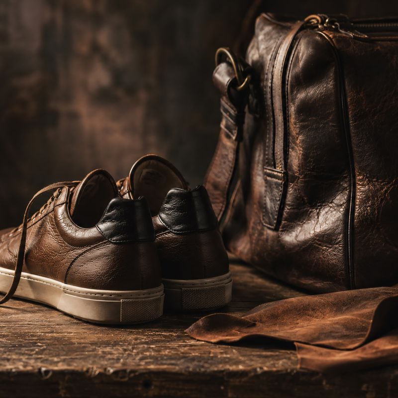Jasperosso brown leather sneakers next to a leather travel bag.